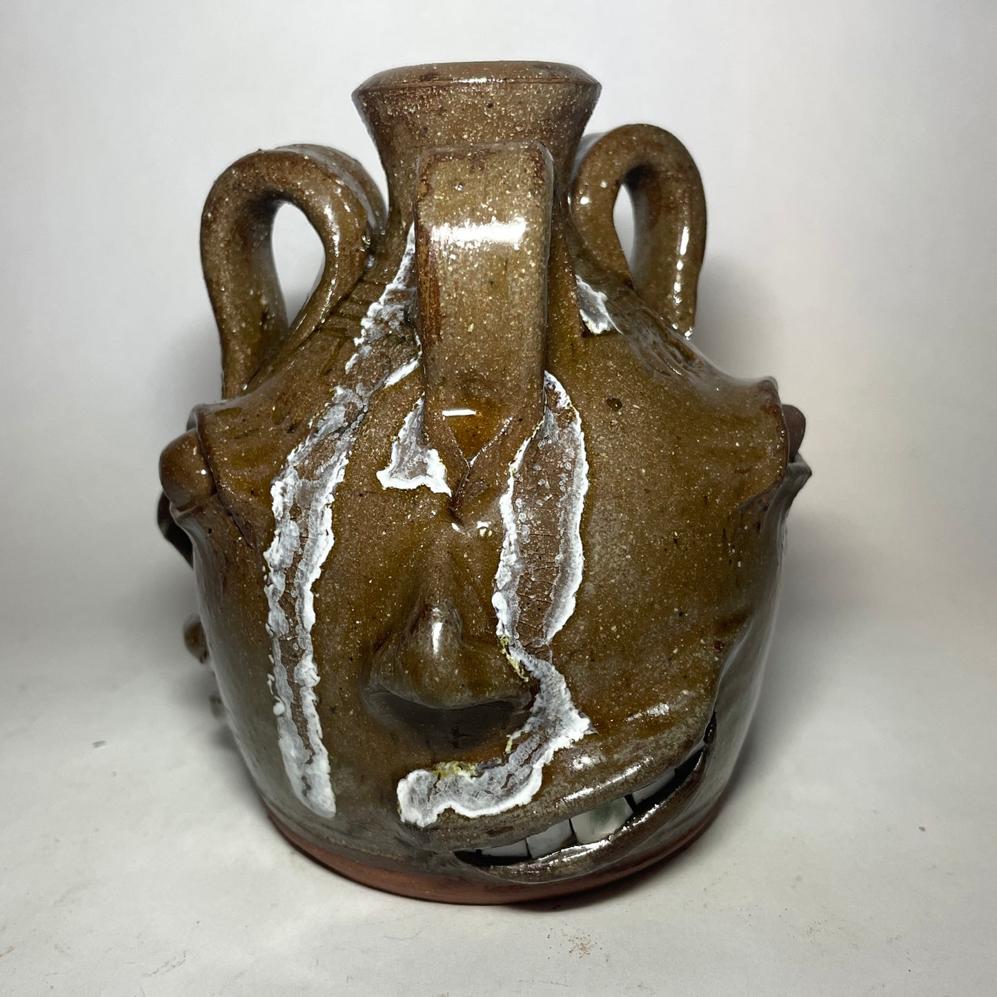 Face 3 of triple face jug. Brown clay, clear glaze. Broken china plate teeth. Glass runs from handle.