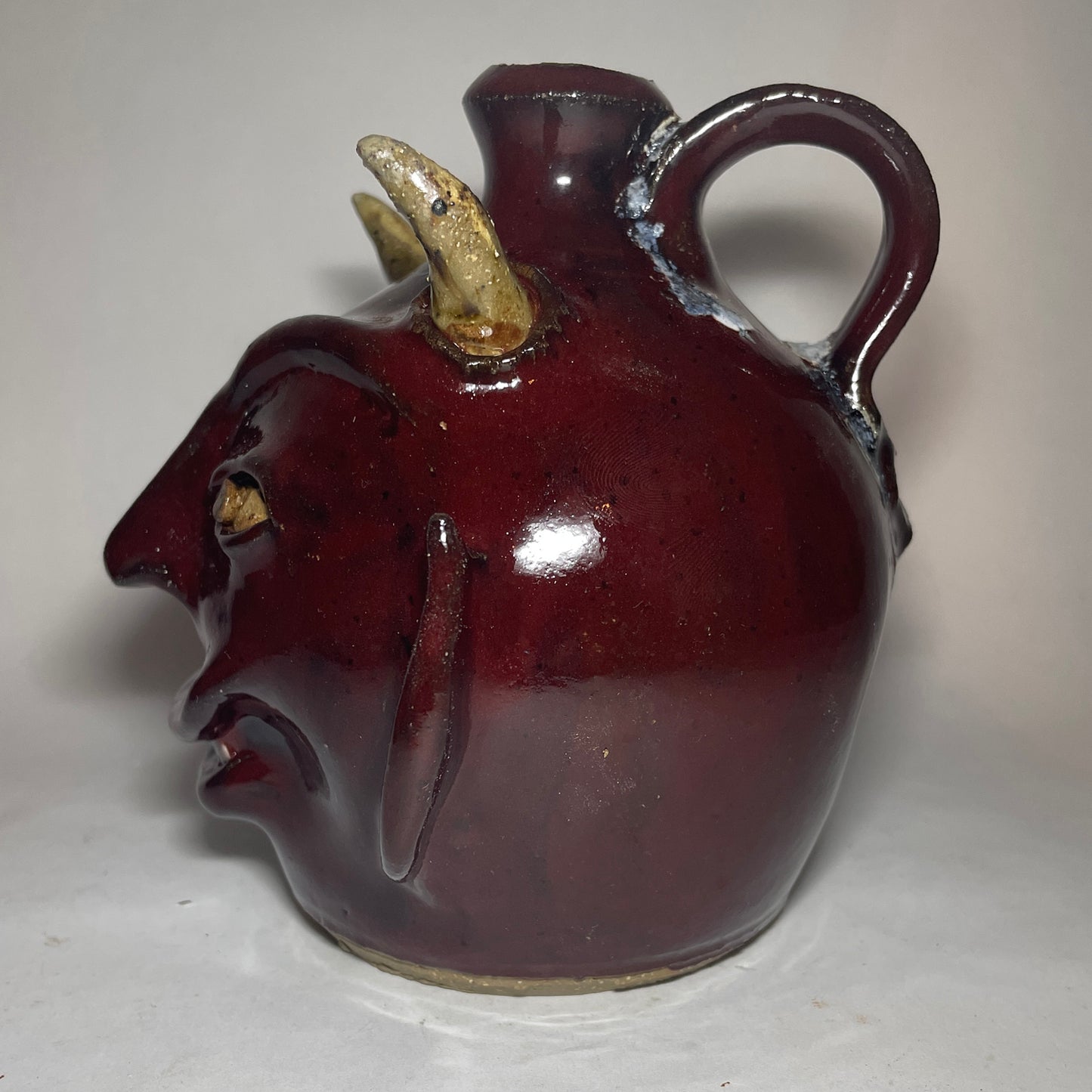Right side view of a devil face jug with red glaze, two horns, and broken china plate teeth.