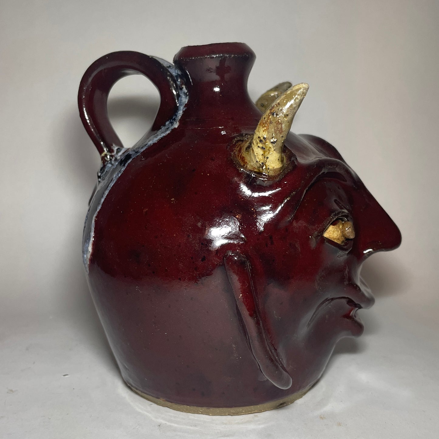 Left side view of a devil face jug with red glaze, two horns, and broken china plate teeth.