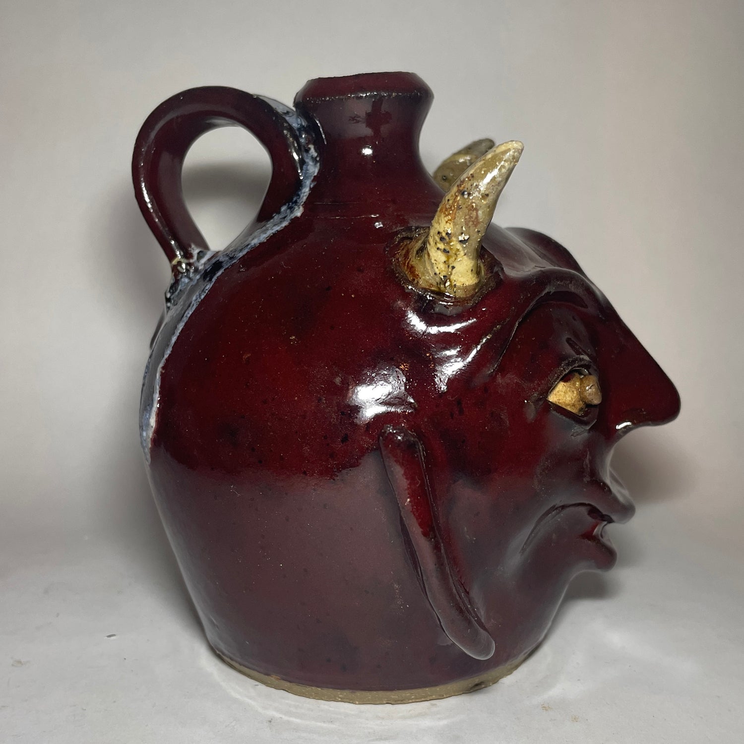 Left side view of a devil face jug with red glaze, two horns, and broken china plate teeth.