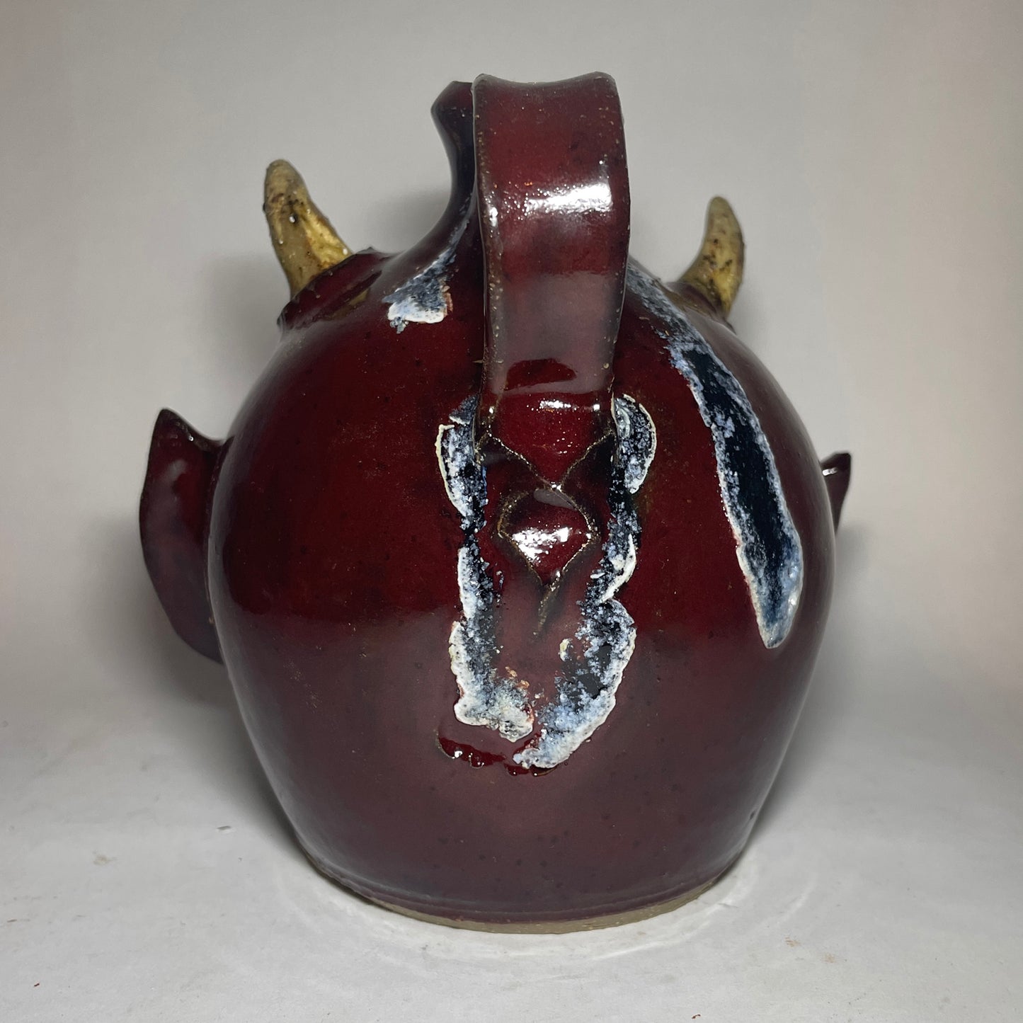 Rear view of a devil face jug with red glaze, two horns, and blue glass runs at the handle.