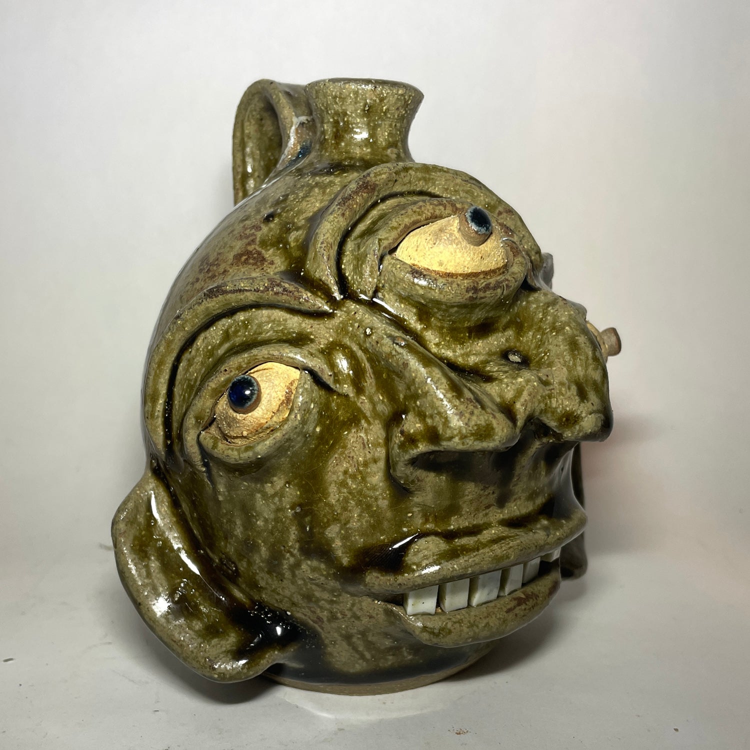 Angled view of a handmade ceramic pot shaped like a stylized, grotesque face with three eyes, two noses merging to one, and a small spout at the top, covered in a textured olive-green and brown glaze.