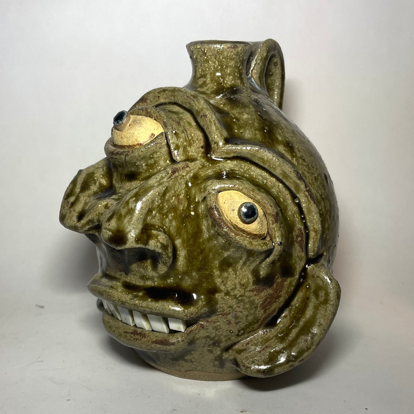 Angled view of a handmade ceramic pot shaped like a stylized, grotesque face with three eyes, two noses merging to one, and a small spout at the top, covered in a textured olive-green and brown glaze.