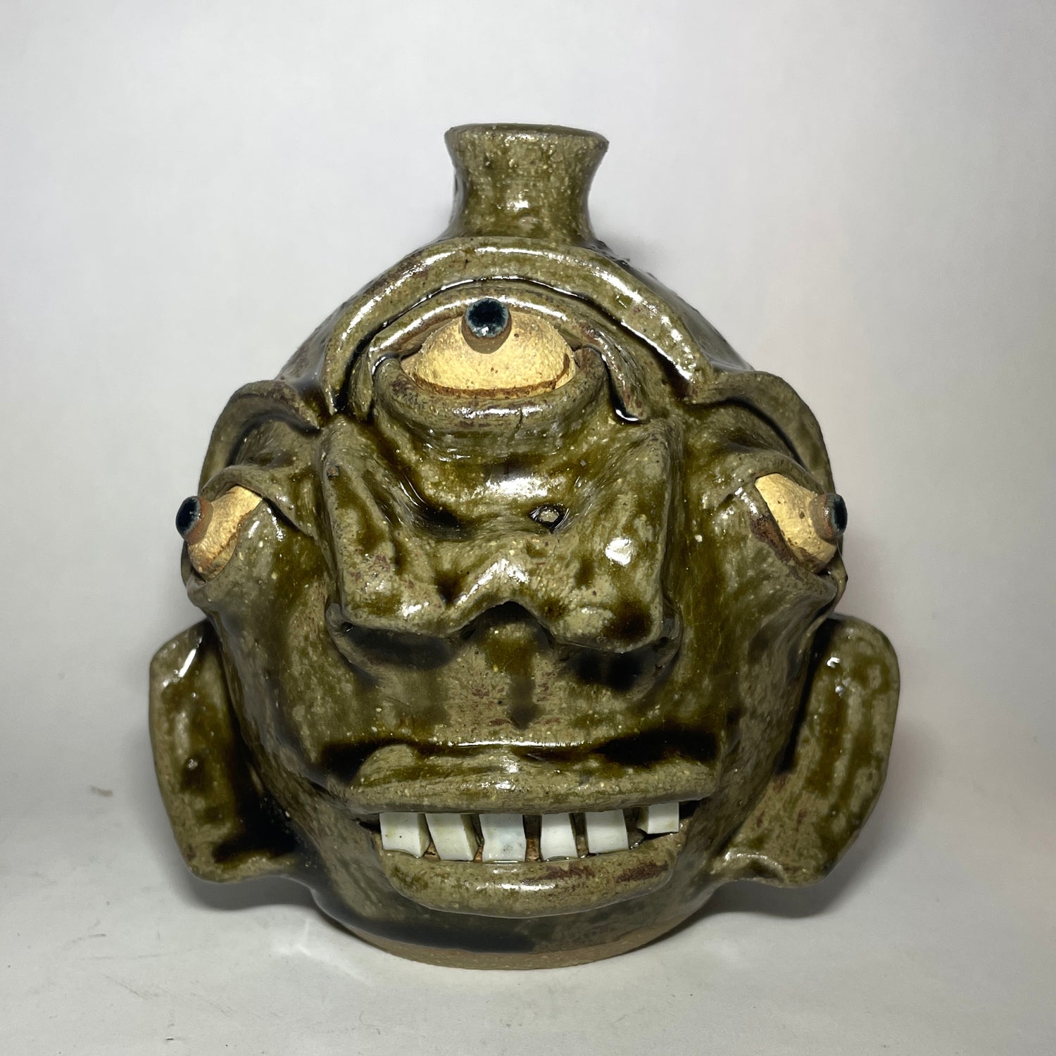 A handmade ceramic pot shaped like a stylized, grotesque face with three eyes, two noses merging to one, and a small spout at the top, covered in a textured olive-green and brown glaze.
