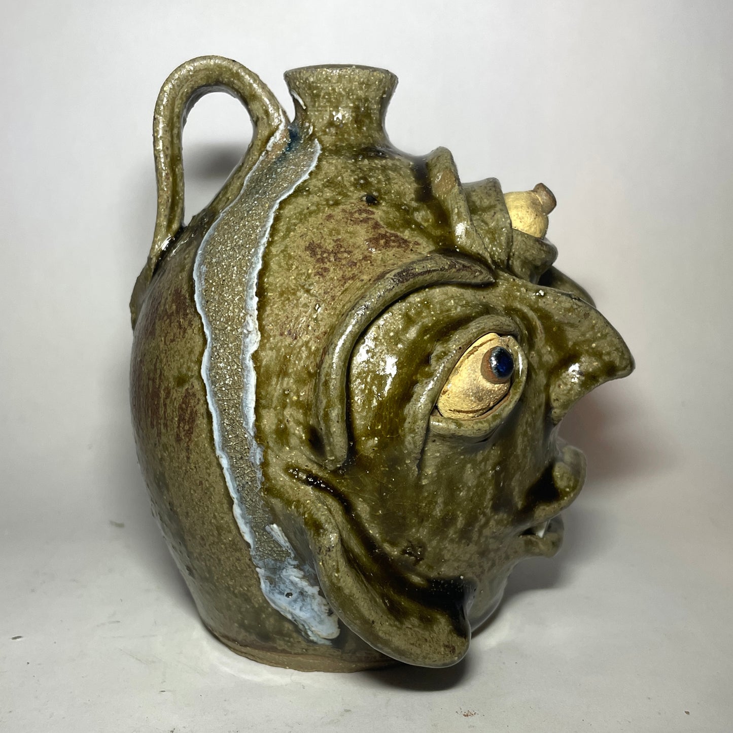 Side view of a handmade ceramic pot shaped like a stylized, grotesque face with three eyes, two noses merging to one, and a small spout at the top, covered in a textured olive-green and brown glaze. Glass run seen at handle.
