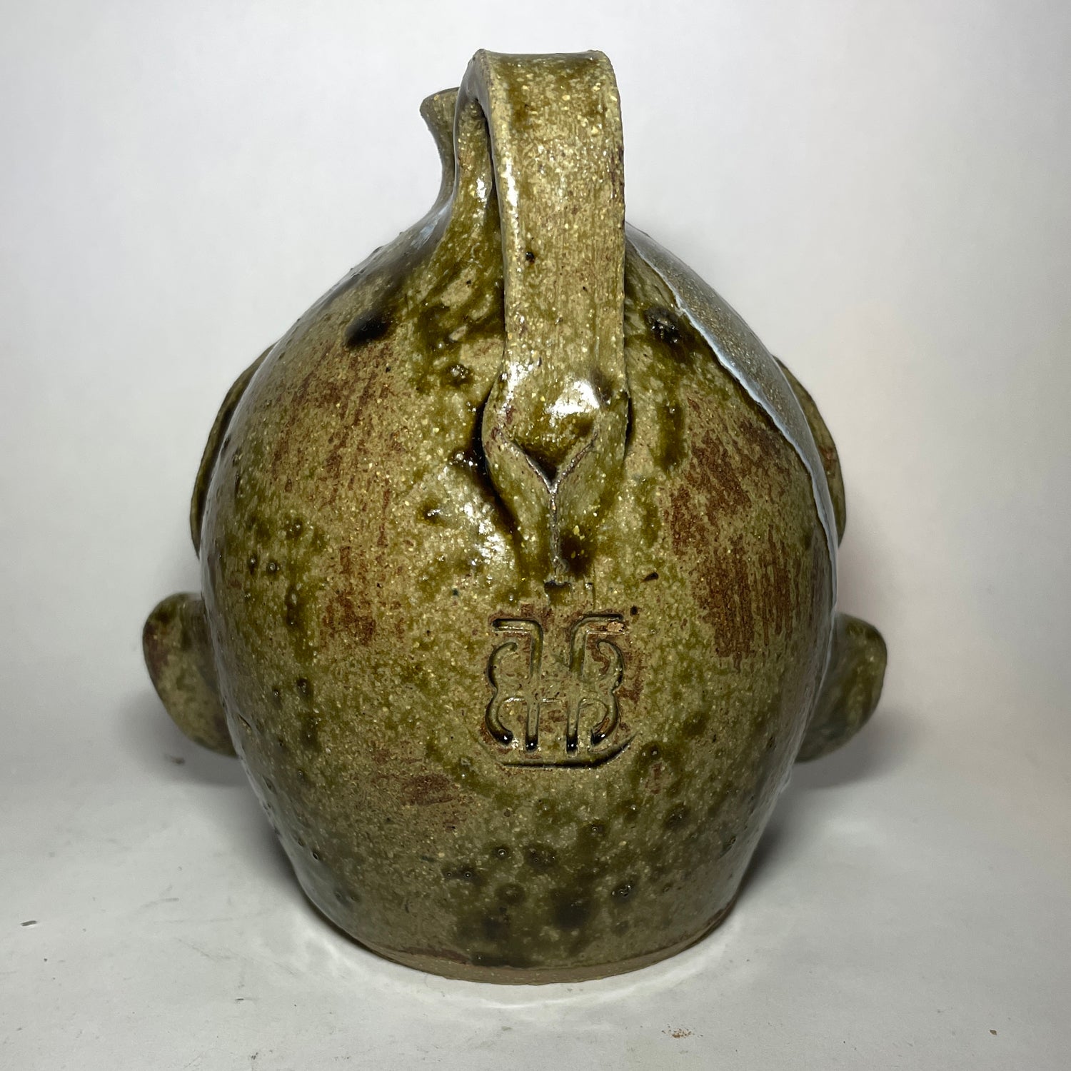 Back view of a handmade ceramic pot shaped like a stylized, grotesque face with three eyes, two noses merging to one, and a small spout at the top, covered in a textured olive-green and brown glaze. Marker mark below handle.