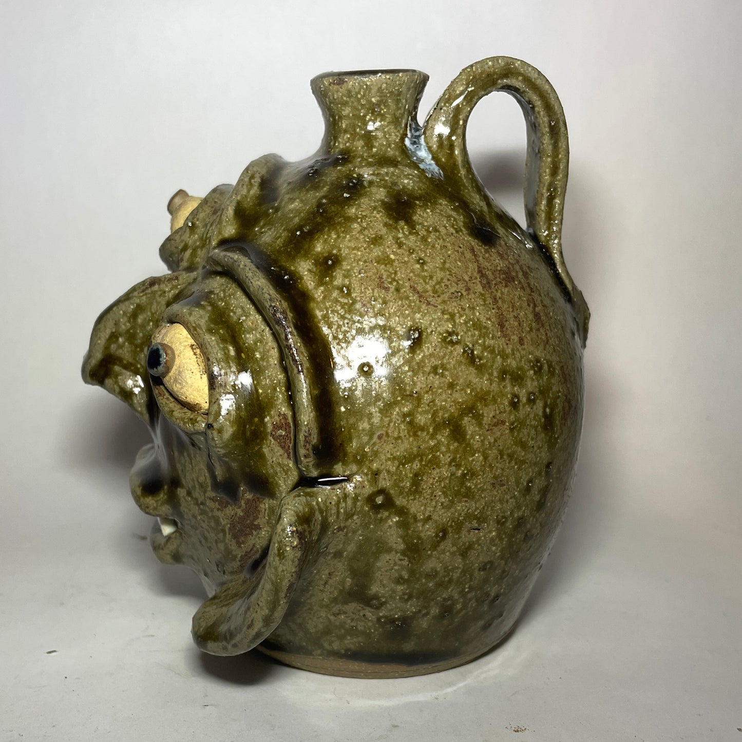 Side view of a handmade ceramic pot shaped like a stylized, grotesque face with three eyes, two noses merging to one, and a small spout at the top, covered in a textured olive-green and brown glaze.